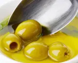 Kilka zielonych oliwek w złocistej oliwie, łyżka zanurzona w płynie. Oliwa z oliwek kcal - zdrowie i smak.