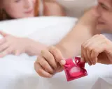 Mężczyzna otwiera prezerwatywę w czerwonym opakowaniu, przygotowując się do intymnych chwil. Bezpieczeństwo przede wszystkim, nawet w najbardziej namiętnym momencie.
