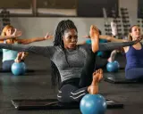 Frauen in einem Kurs, der Pilates Krafttraining macht, sitzen auf Matten mit kleinen blauen Bällen unter ihren Füßen.