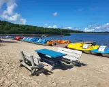Słoneczny dzień nad jeziorem Sarbsko. Rząd kolorowych rowerów wodnych czeka na plaży, obok drewniane ławki i stół.