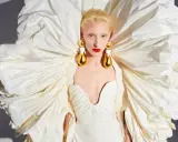 A model wears a dramatic white gown with a voluminous, ruffled cape, showcasing Daniel Roseberry's avant-garde design and striking gold teardrop earrings.