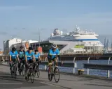Fünf Radfahrer in blauen Trikots trainieren für einen Mini-Triathlon am Hafen, ein großes Kreuzfahrtschiff im Hintergrund.