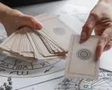 Dłonie trzymają talię kart, a jedna z nich wybiera kartę z symbolem paz denarow. W tle widoczny jest wykres astrologiczny.