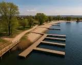 Drewniane pomosty w kształcie litery L i proste, prowadzące do wody, na piaszczystej plaży. W tle zielone drzewa i zabudowania. Idealne miejsce na wypoczynek nad wodą, jak na przylasku ruskim.