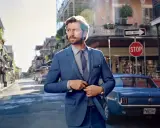 Man in a sharp blue suit, ready for a business cocktail, stands on a street with a vintage car and classic architecture.