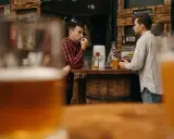 Dwóch mężczyzn rozmawia przy barze, zastanawiając się, ile gram alkoholu ma piwo. Na blacie stoją kufle i piwo.