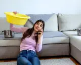Mujer preocupada llama por teléfono mientras sostiene un barreño amarillo para recoger agua de las goteras en casa. Ollas esperan en el sofá.