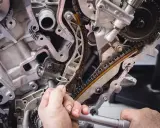 Mechanik dokręca śrubę przy rozrządzie silnika. Widać fragmenty łańcucha rozrządu i metalowe elementy, które mogą być częścią poduszki silnika.