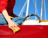 Main d'une personne lavant une coque de bateau rouge avec une éponge jaune.