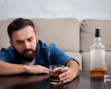 Mężczyzna z zarostem patrzy na szklankę z alkoholem, widoczne objawy uzależnienia. Obok pusta butelka i kolejna szklanka.