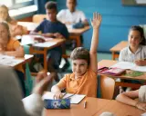 Chłopiec z podniesioną ręką czeka na ocenianie odpowiedzi. Inne dzieci pracują przy ławkach.