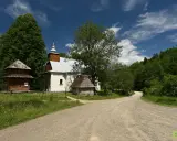 Cerkiew w Łopience, drewniana chata i kamienna przybudówka na tle zieleni i błękitnego nieba z chmurami.