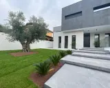 Moderna casa con fachadas color gris y blanco, escaleras de mármol y un olivo centenario en el jardín.
