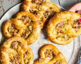 Herzhafte Brezeln mit Lauch und Speck, die an Flammkuchen erinnern, liegen auf einem Blech. Eine Hand greift nach einer Brezel.
