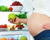 Gravid kvinna tar ett grönt äpple ur kylen fylld med färsk frukt och grönsaker. Hon njuter av hälsosam mat under sin soja graviditet.