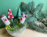 Uroczy stroik DIY z korkowych ludzików, grzybków i choinek w szklanej misie, ozdobiony gałązkami świerku i lampkami.
