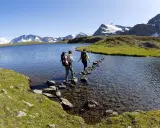 Due escursionisti attraversano il lago di Ceresole su un sentiero di pietre, con montagne innevate sullo sfondo.