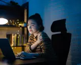 Une jeune femme, en pleine **mise en place télétravail**, ressent une douleur à l'épaule devant son ordinateur, éclairée par une lampe de bureau.