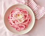 En skål med rosa pasta, toppad med pinjenötter och riven ost. En randig duk i rosa och vitt ligger bredvid.