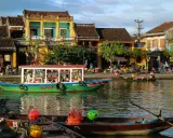 Båtar flyter på floden i Hoi An, Vietnam, med färgglada hus och folk som promenerar längs kajen.