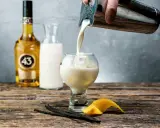 En hand häller en krämig dryck från en shaker i ett glas med is. Bredvid står en flaska Licor 43, en karaff med mjölk, vaniljstänger och citronskal.