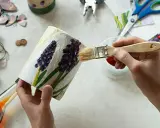 Ręce malują pędzlem ozdobiony kwiatami pojemnik, wokół materiały do decoupage.