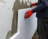 Pracownik w czerwonych rękawicach montuje styropian na elewacji domu parterowego.