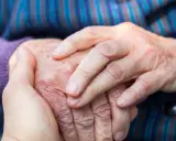 Dłonie młodszej osoby obejmują dłoń starszej, symbolizując wsparcie w obliczu choroby Alzheimera i jej przyczyn.