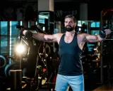 Mann mit Bart trainiert im Fitnessstudio, macht seitheben sitzend mit Hanteln.