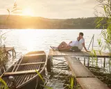 Para, gdzie na Mazury we dwoje? Para siedzi na pomoście, łódka przy brzegu, zachód słońca.