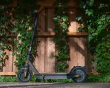 Trottinette électrique noire, prête à l'aventure, avec une puissance impressionnante, posée devant un mur de bois couvert de végétation.