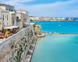 Vista del lungomare di Otranto centro, con edifici bianchi, un ristorante all'aperto e il mare azzurro cristallino.