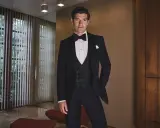 A man in a sharp black tie tuxedo, ready for a wedding. He stands confidently in a stylish room with large windows and wood paneling.
