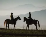 Due cavalieri in silhouette godono di una **passeggiata a cavallo in Lombardia** al tramonto, con montagne nebbiose all'orizzonte.