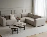 En snygg hörnsoffa i ljusbeige tyg, med kuddar och ett runt soffbord med blommor.