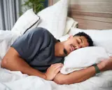 Ein Mann schläft tief und fest, um sein HRV zu verbessern. Er liegt auf einem weißen Bett mit vielen Kissen.