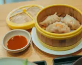 Parujące pierogi gyoza w bambusowym parowniku, obok miseczka z sosem.