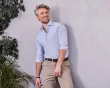 Uśmiechnięty mężczyzna w niebieskiej koszuli w paski i beżowych chinosach na wesele, oparty o ścianę, otoczony zielenią.