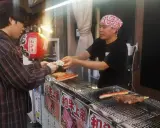 Un cliente paga por comida callejera japonesa: brochetas de carne y surimi a la parrilla en un puesto.