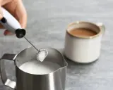 Hand hält Milchaufschäumer über Metallkännchen mit Milch. Perfekt für leckere Milchschaum Rezepte.