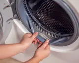 Ręce podnoszą gumową uszczelkę drzwi pralki, odsłaniając widoczny szlam w pralce i brud.
