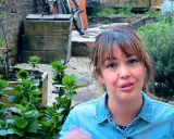 Mujer sonriente en un jardín con plantas verdes y macetas.