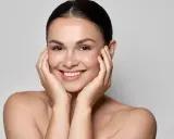Uśmiechnięta kobieta z idealną, lśniącą cerą typu "glass skin", delikatnie dotykająca policzków dłońmi.