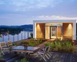 Moderner dachgarten auf einem Flachdach mit Blick auf einen See. Holzdeck, Sitzgelegenheiten und üppige Bepflanzung laden zum Entspannen ein.