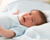 Płaczące niemowlę w niebieskim body leżące na białym kocyku.