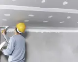 Pracownik w żółtym kasku nakłada masę szpachlową na sufit, przygotowując go do montażu. To kluczowy etap, by wiedzieć, jak przykleić styropian do sufitu.