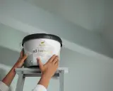 Händer håller en hink med Alcro ad.harmoni takfärg på en stege, redo att måla tak utan dropp.