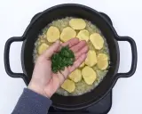 Manos añaden perejil picado a patatas en rodajas y cebolla en una sartén, preparando patatas en salsa verde.
