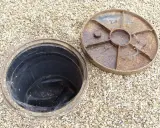 Inspection d'une canalisation bouchée sous la maison. Le couvercle rouillé est retiré, révélant le conduit noir.