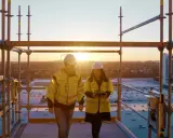 Två byggarbetare i varselkläder inspekterar ett bygge vid solnedgången. De diskuterar säkerhetslösningar, som brandklass EI60.